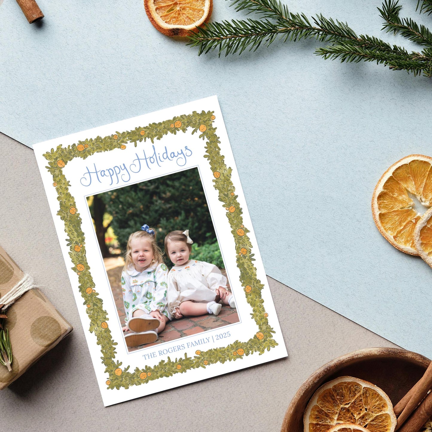 A holiday card with a photo of two young girls sitting on a brick path, surrounded by holiday decorations such as pine branches, dried orange slices, and cinnamon sticks.