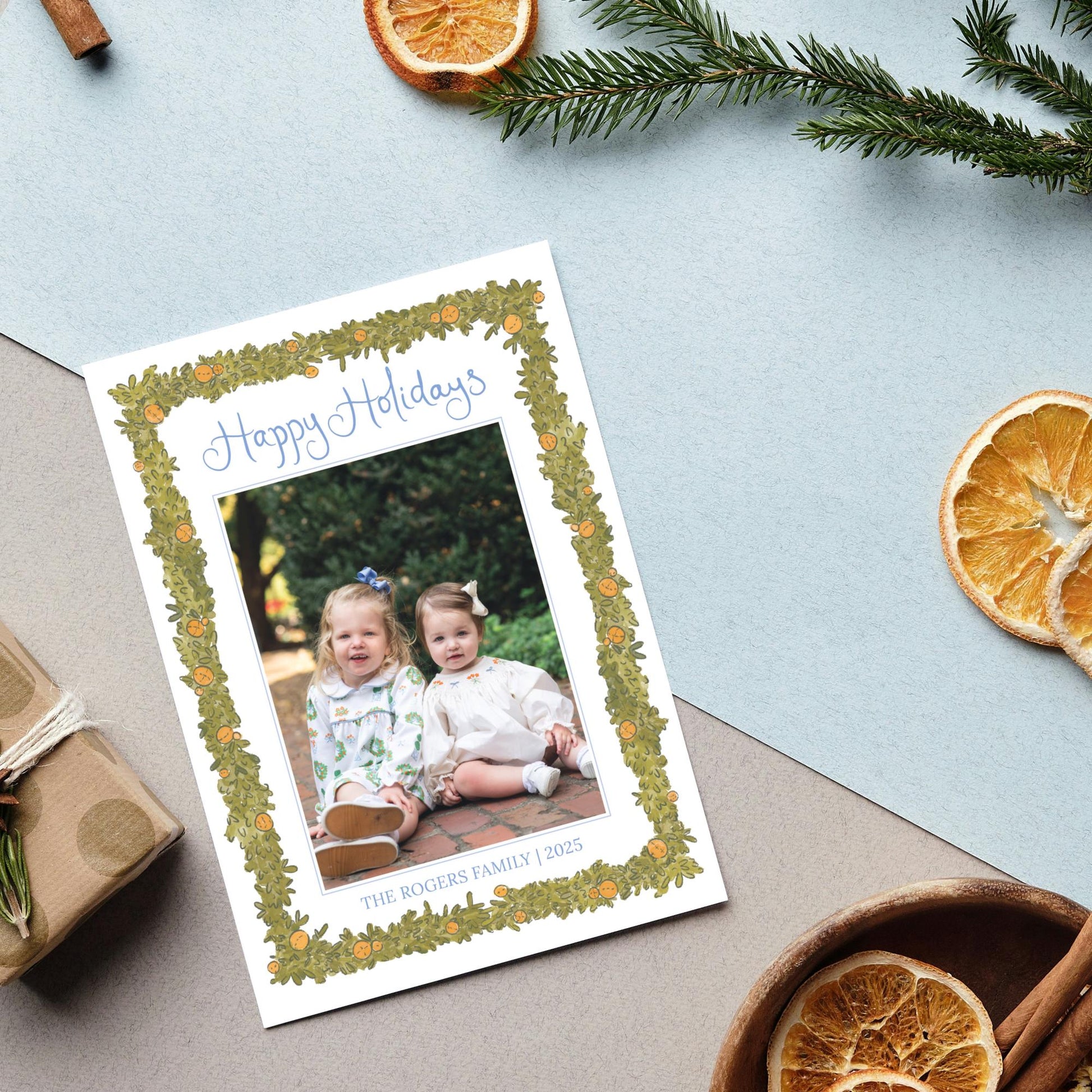 A holiday card with a photo of two young girls sitting on a brick path, surrounded by holiday decorations such as pine branches, dried orange slices, and cinnamon sticks.