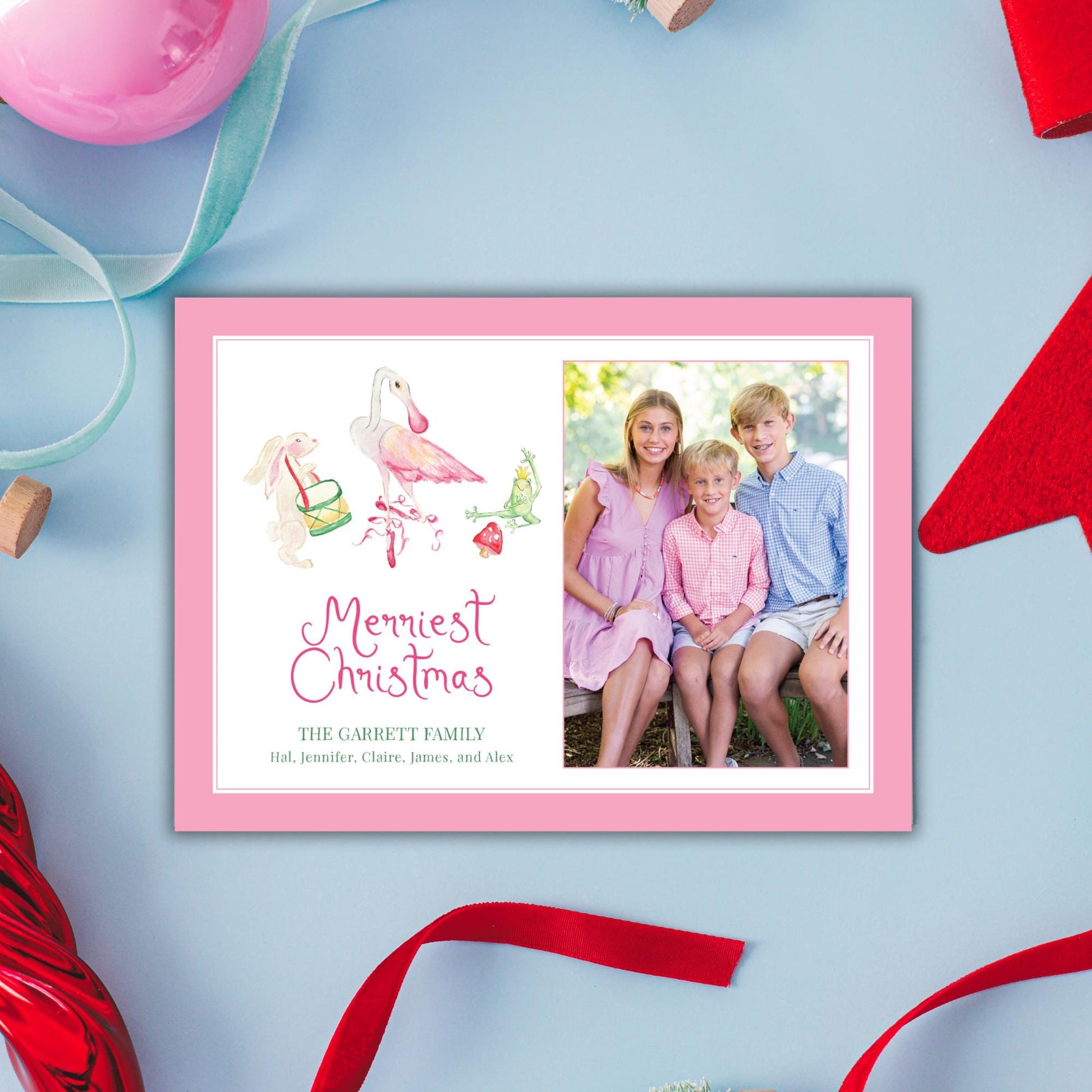 A pink and white Christmas card featuring a family photo of three children and two adults, with a watercolor illustration of a pink flamingo and a red ribbon.