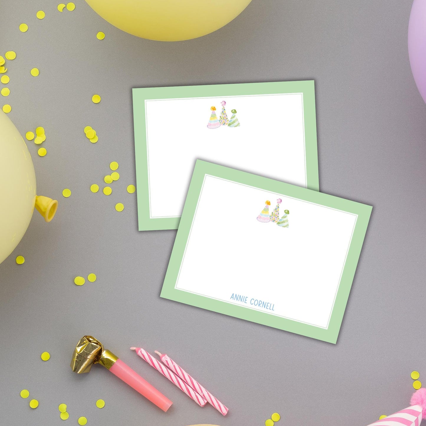 two green-framed cards with illustrations of party hats, along with a pink candle holder, confetti, and a gold foil object.