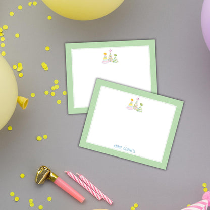 two green-framed cards with illustrations of party hats, along with a pink candle holder, confetti, and a gold foil object.