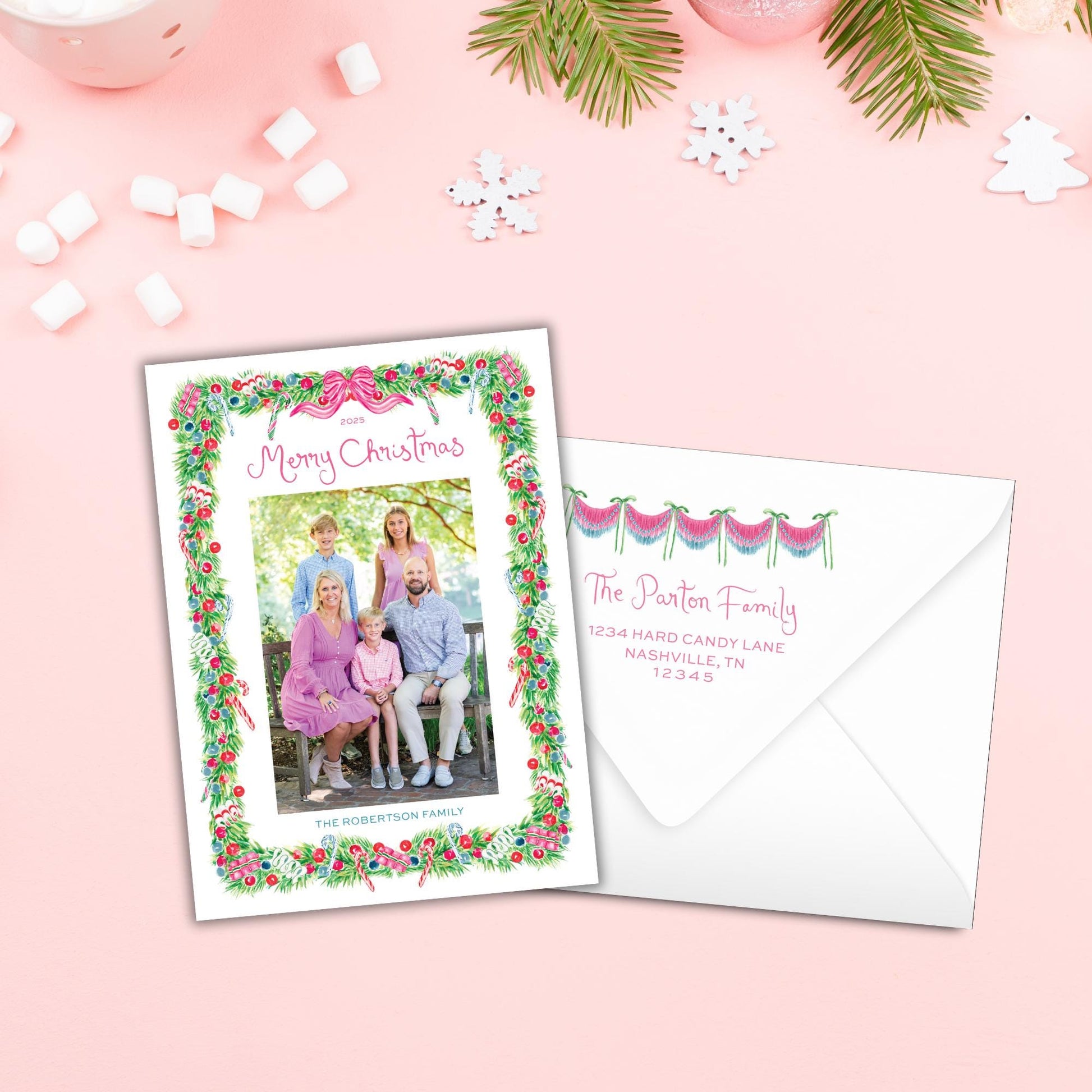 A Christmas card with a family photo and a white envelope with a personalized address, both placed on a pink background with snowflakes and other holiday decorations.