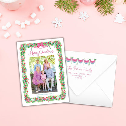 A Christmas card with a family photo and a white envelope with a personalized address, both placed on a pink background with snowflakes and other holiday decorations.