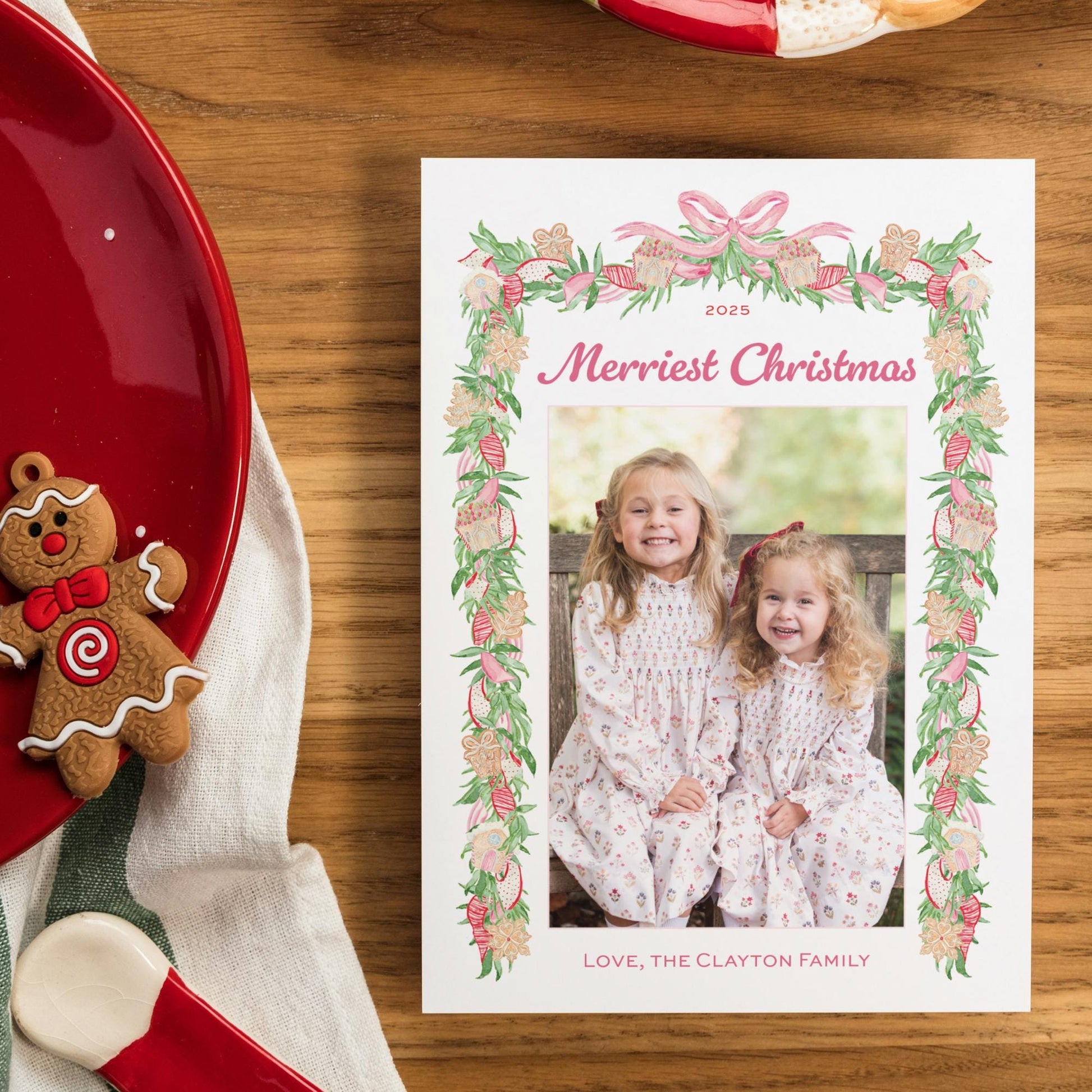 A 2015 Christmas card with a photo of two young girls, surrounded by a floral border, sits on a wooden table next to a red plate with a gingerbread man decoration.