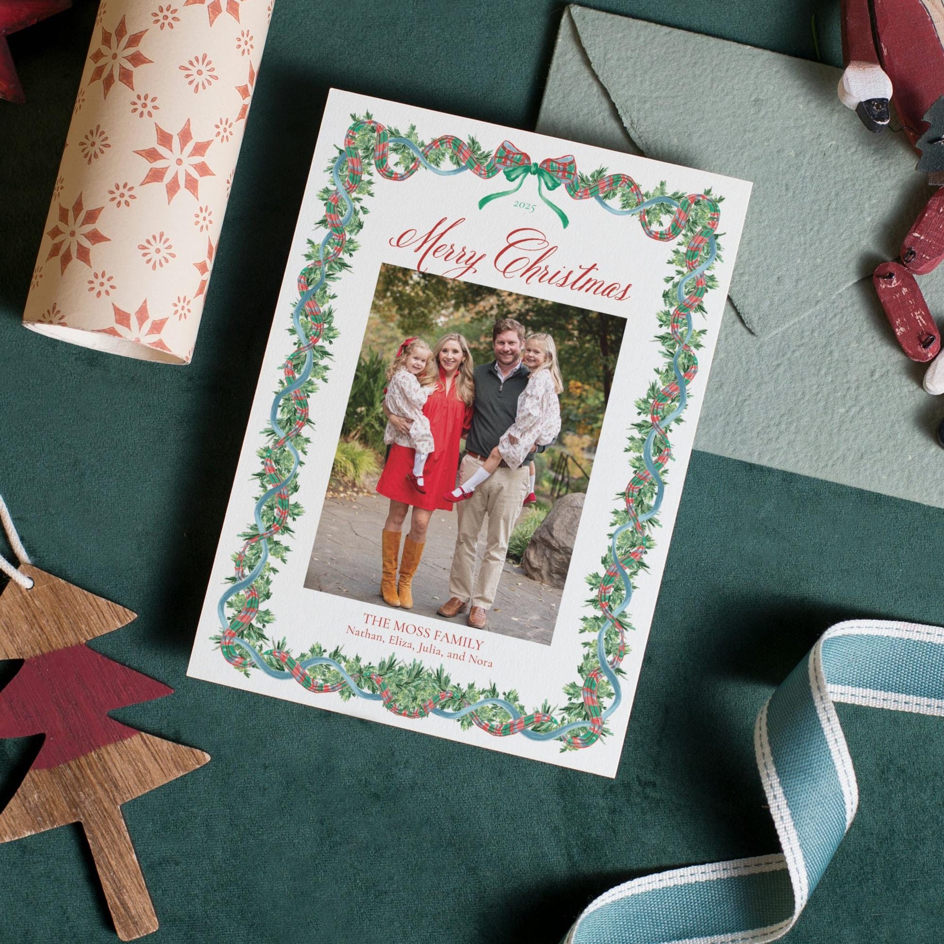 A Christmas card with a family photo of a man, woman, and two children, surrounded by a festive border of holly and berries. The card is placed on a green surface, with other holiday decorations and items visible in the background.