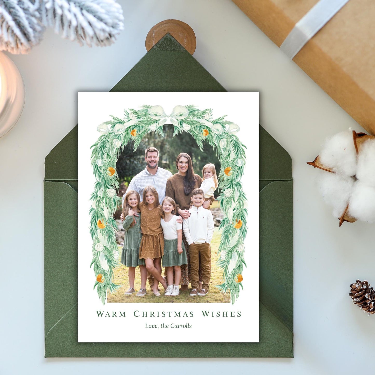 A family portrait of a man, woman, and three children, surrounded by a wreath of greenery and orange flowers, placed inside a green envelope.