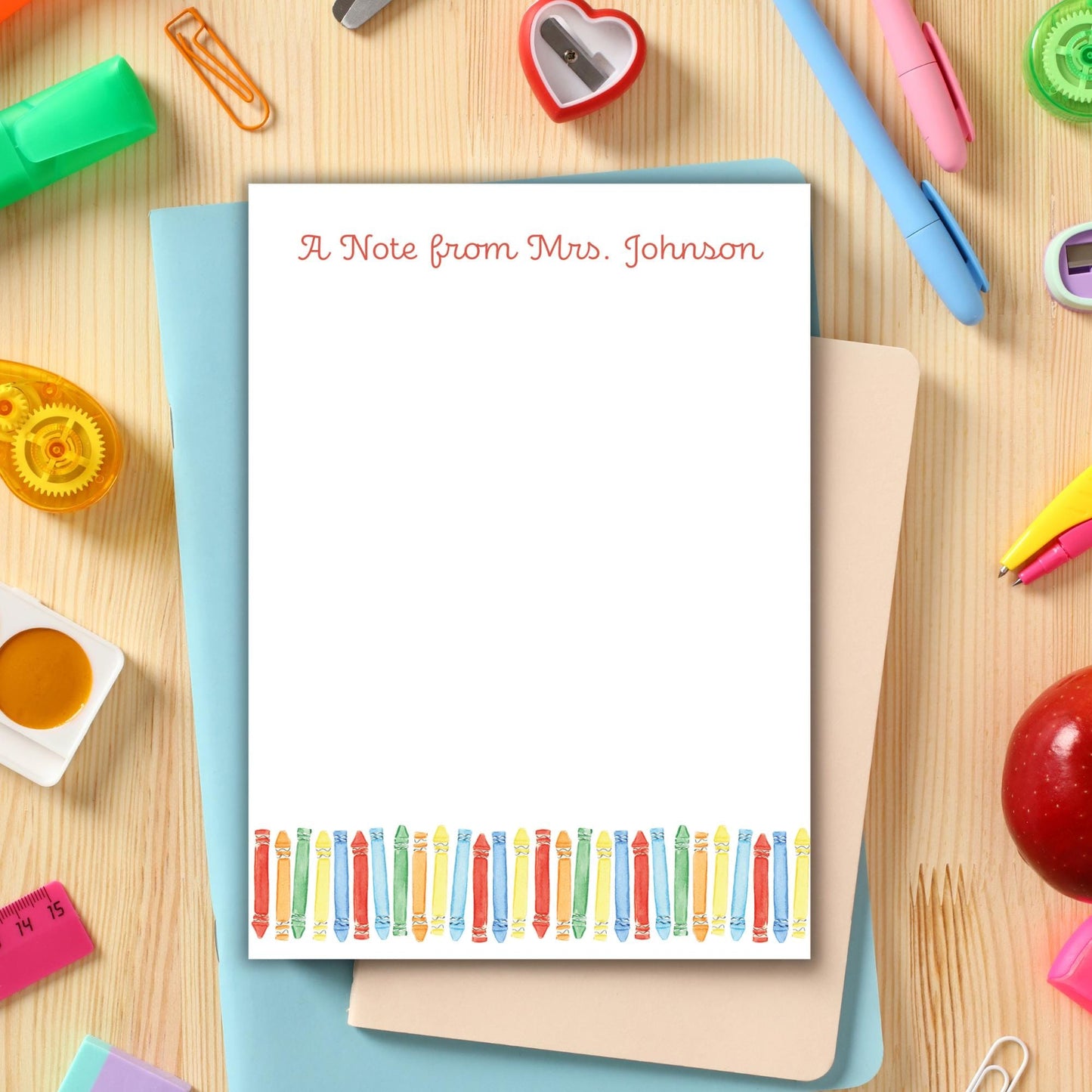 A blank note card with a personalized message is placed on a wooden surface surrounded by various school supplies, including pens, pencils, and a heart-shaped object.
