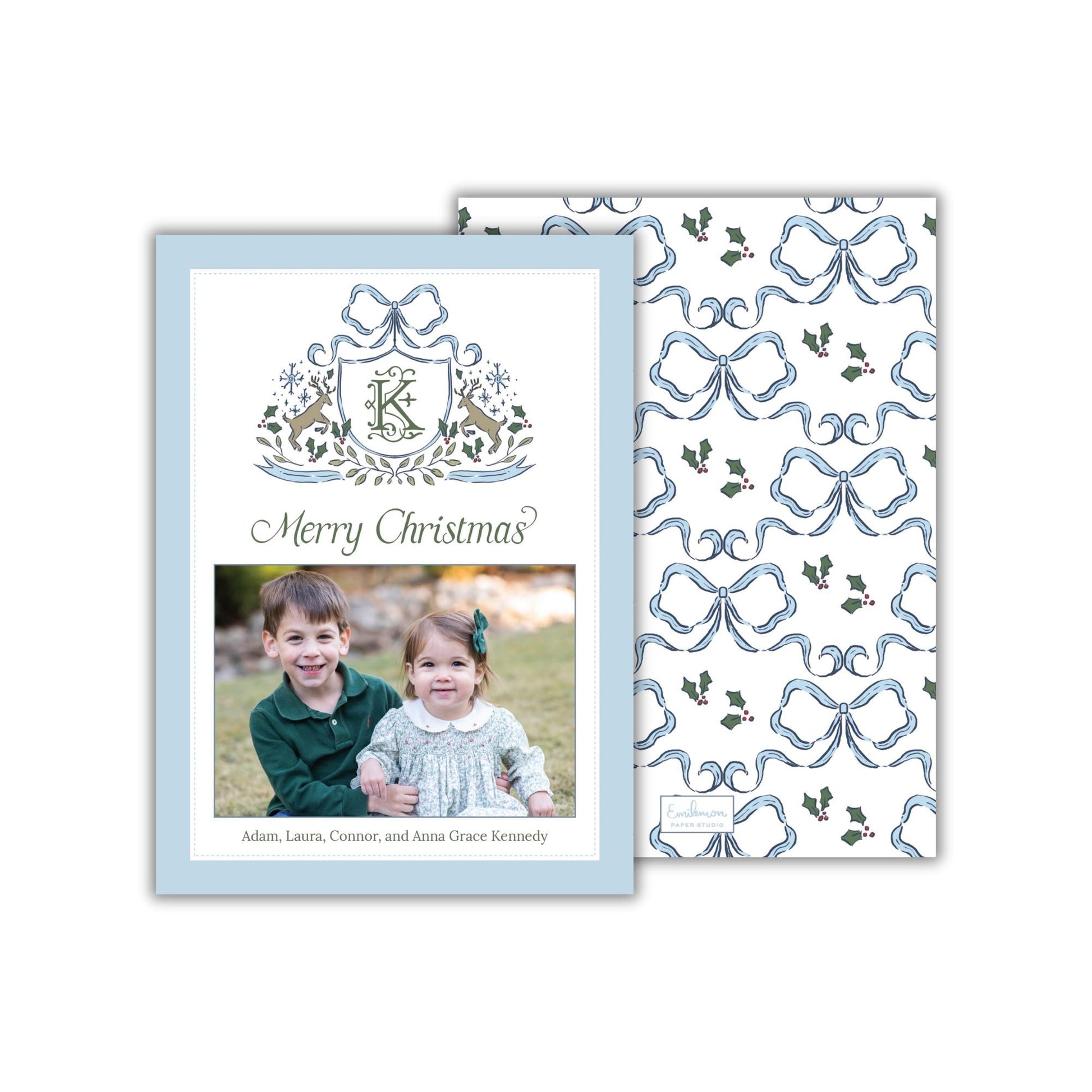 A Christmas card featuring a photo of two children, likely siblings, smiling together. The card has a blue and white color scheme with a repeating pattern of blue bows and holly leaves. The text on the card reads &quot;Merry Christmas?&quot; and includes the names &quot;Adam, Laura,Connor, and Anna Grace Kennedy&quot;.
