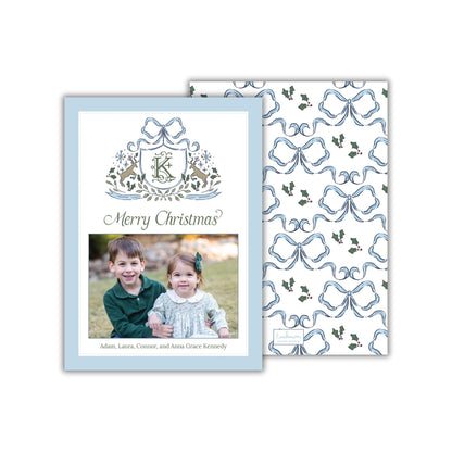 A Christmas card featuring a photo of two children, likely siblings, smiling together. The card has a blue and white color scheme with a repeating pattern of blue bows and holly leaves. The text on the card reads "Merry Christmas?" and includes the names "Adam, Laura,Connor, and Anna Grace Kennedy".