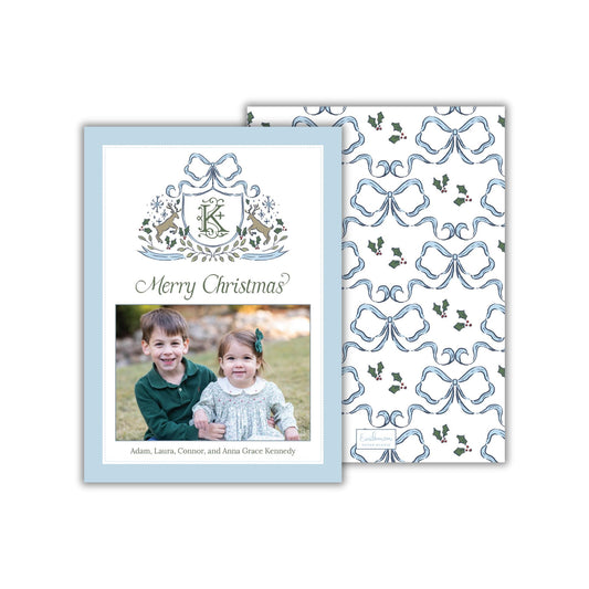 A Christmas card featuring a photo of two children, likely siblings, smiling together. The card has a blue and white color scheme with a repeating pattern of blue bows and holly leaves. The text on the card reads &quot;Merry Christmas?&quot; and includes the names &quot;Adam, Laura,Connor, and Anna Grace Kennedy&quot;.