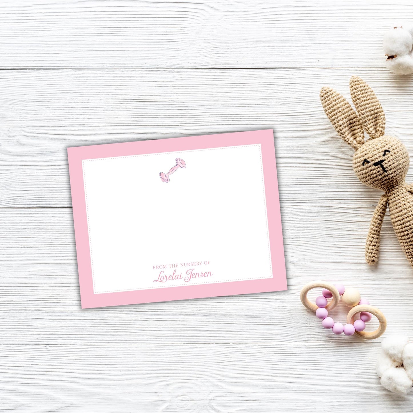 A pink greeting card with a blank space for a message, placed on a white wooden surface next to a crocheted stuffed rabbit toy and some baby toys.