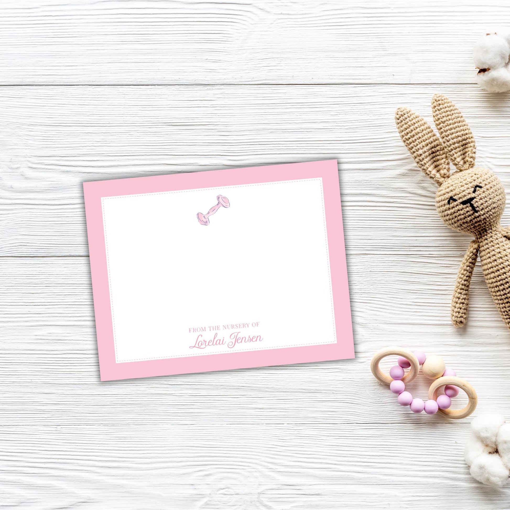 A pink greeting card with a blank space for a message, placed on a white wooden surface next to a crocheted stuffed rabbit toy and some baby toys.