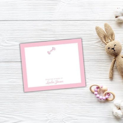 A pink greeting card with a blank space for a message, placed on a white wooden surface next to a crocheted stuffed rabbit toy and some baby toys.