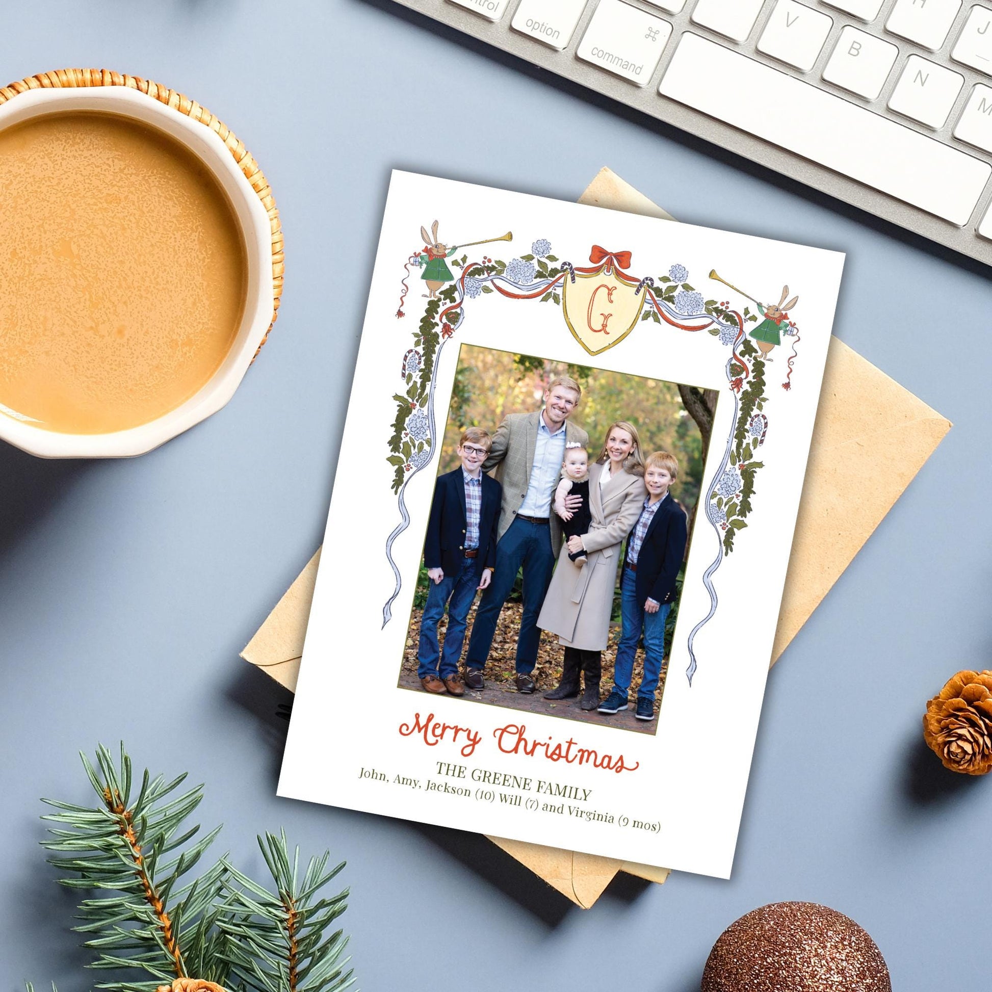 A Christmas card with a family photo is placed on a desk next to a keyboard, a cup of coffee, and some pine branches.
