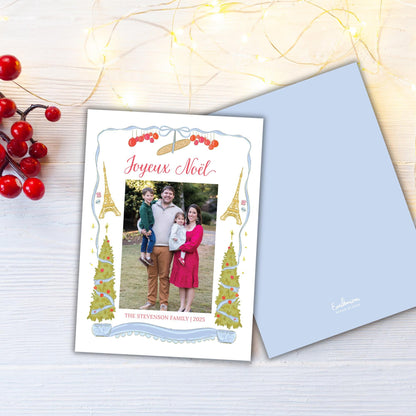 A family photo is displayed on a card with the text "Joyeux Noel" and "The Steveston Family 2015" printed on it. The card is placed on a wooden surface, with a string of lights and red berries in the background.