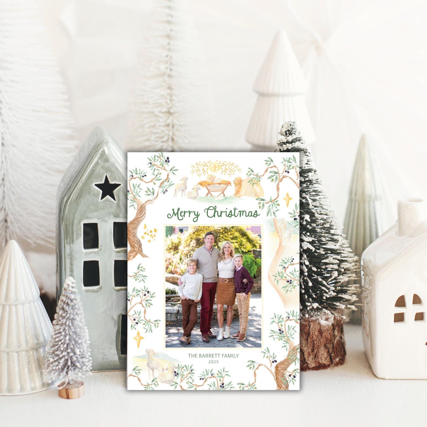 A family photo is displayed in front of a Christmas card with a festive design, surrounded by small white ceramic ornaments and a miniature tree.