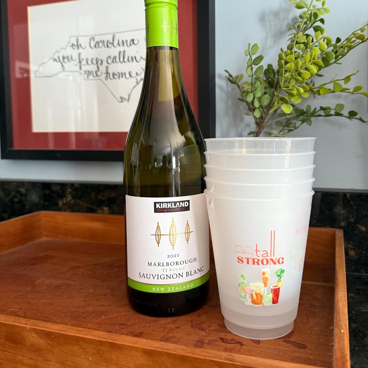 A bottle of wine and a stack of plastic cups are displayed on a wooden tray, with a framed picture of a state in the background.