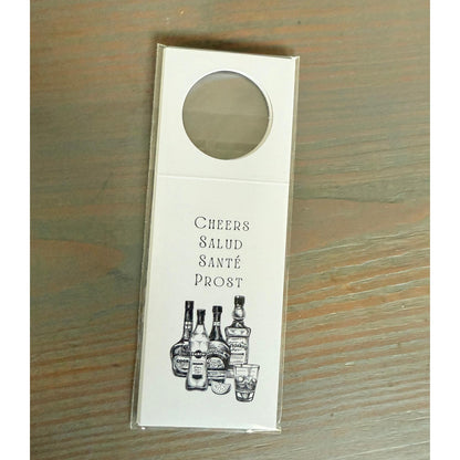 A white door hanger with a circular cutout, featuring illustrations of various alcoholic beverages and the text "Cheers Salud Sábé Prost" printed on it.