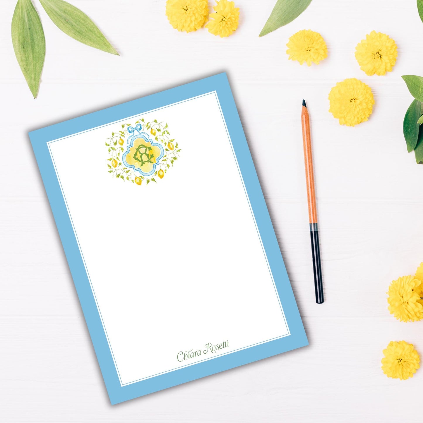 a blue-bordered blank card with a floral design, a pen, and yellow flowers on a white background.
