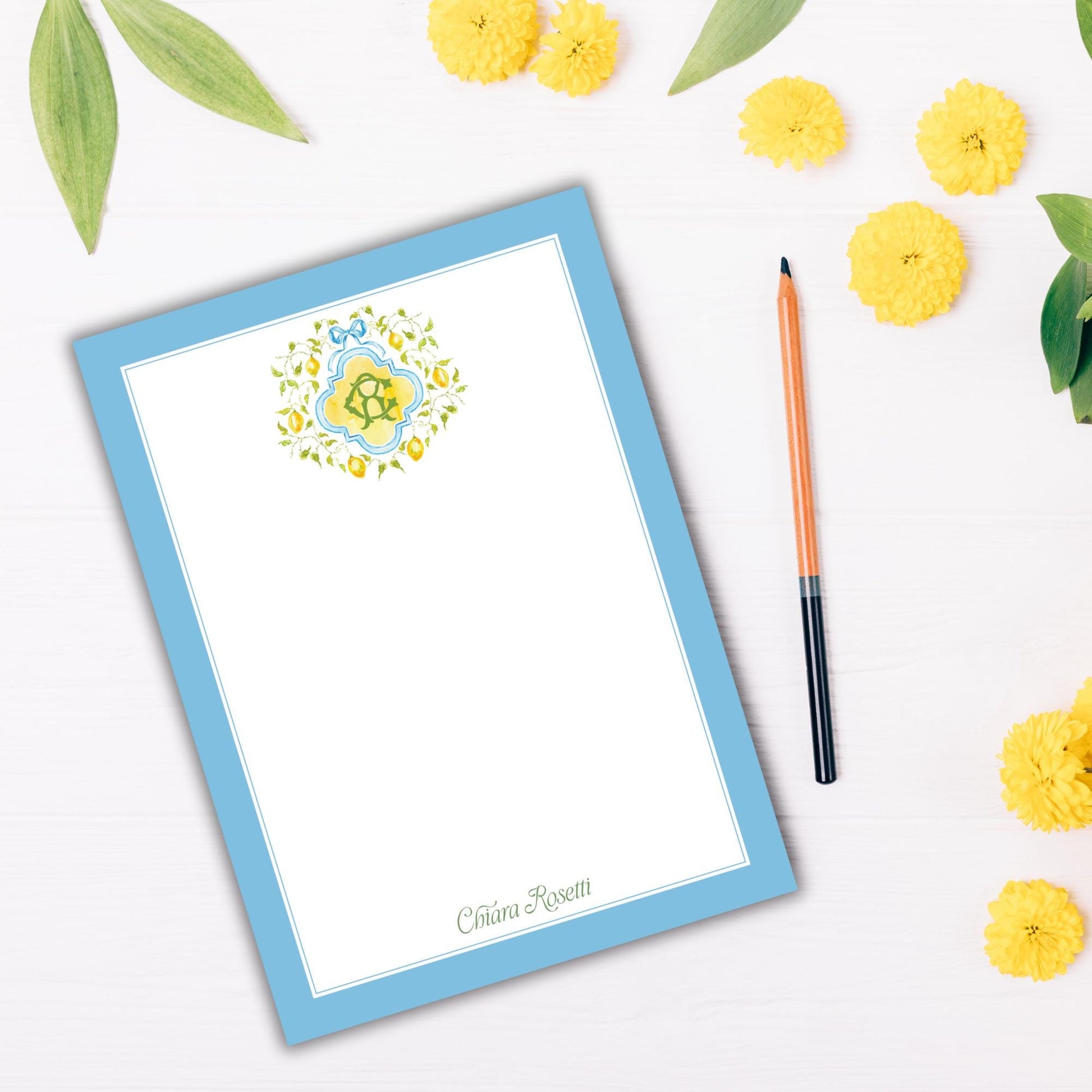 a blue-bordered blank card with a floral design, a pen, and yellow flowers on a white background.