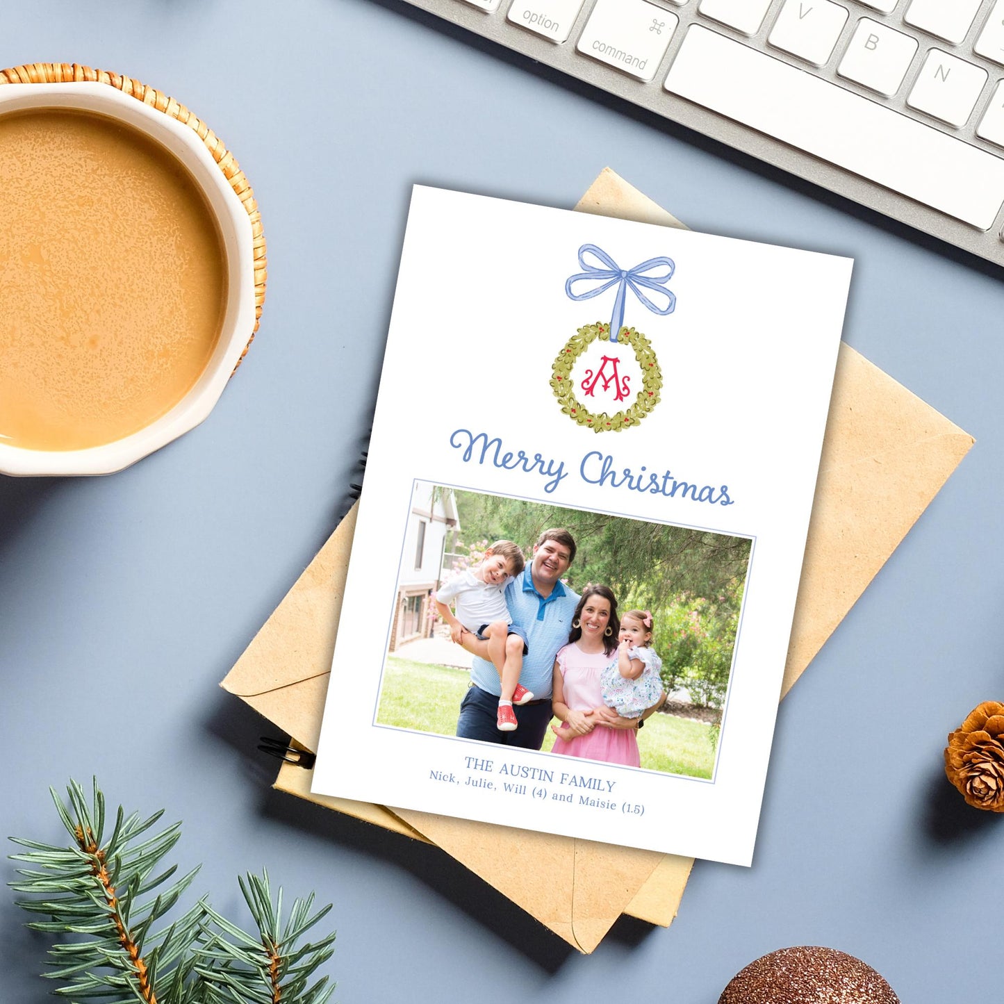 A Christmas card with a photo of a family, along with the text &quot;Merry Christmas&quot; and &quot;The Austin Family&quot; printed on it, placed on a desk next to a keyboard, a mug, and some pine branches.