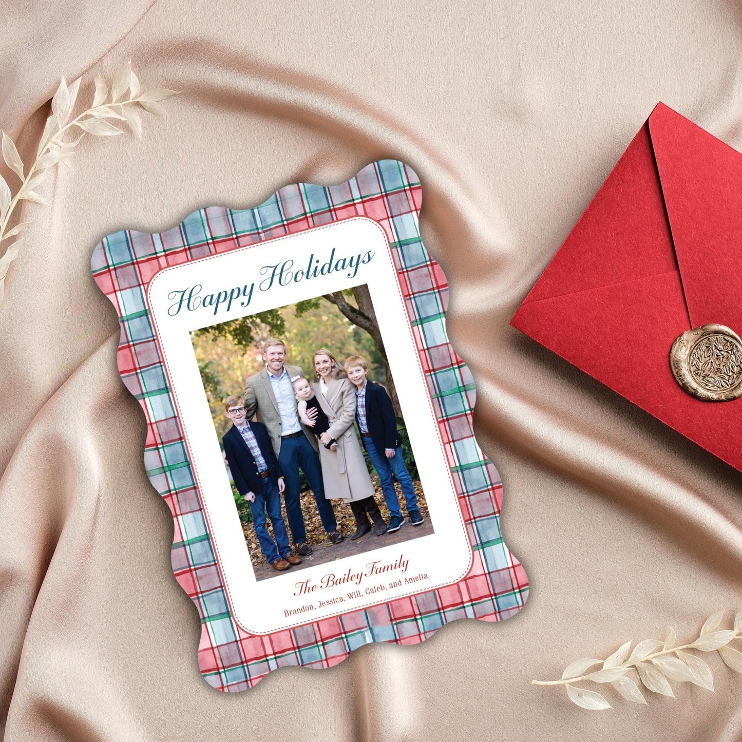 A family photo of five people, likely a family, is displayed on a plaid-patterned card with the text &quot;Happy Holidays&quot; and &quot;The Bailey family&quot; written on it. The card is placed on a pink fabric, and there is a red envelope and some leaves in the background.