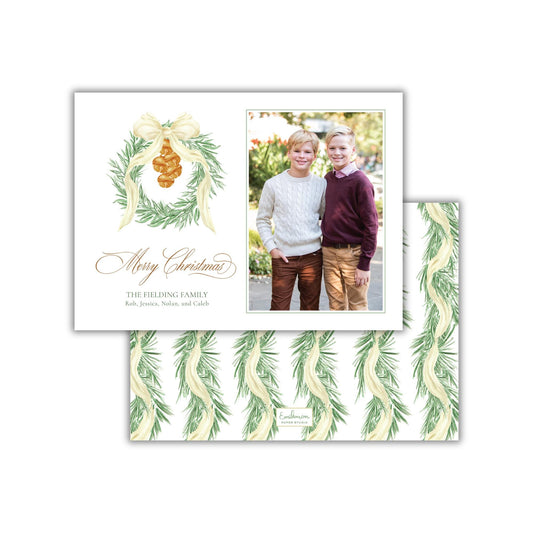 A Christmas card featuring two young boys posing together in front of a wreath. The card has a festive design with a bow and greenery, and includes a photograph of the two boys.