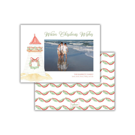 A Christmas card featuring a lighthouse on the beach with three people standing in front of it. The card has a photo of the family in the background.