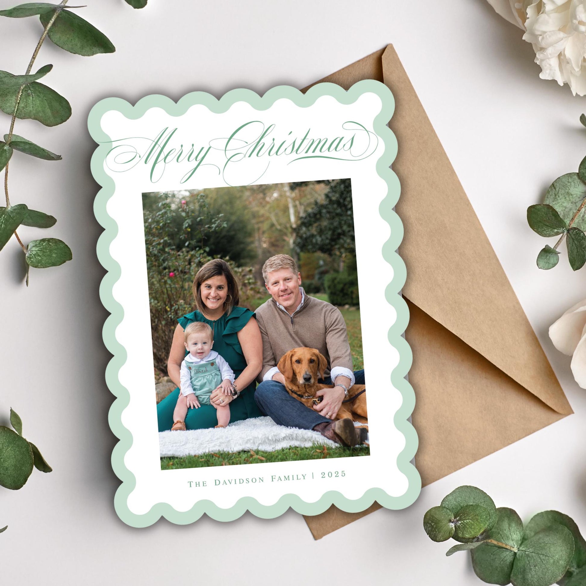 A family photo of a man, woman, and child sitting together on a blanket in a garden setting, with a dog nearby. The photo is framed in a scalloped edge and placed on a brown envelope, surrounded by greenery.