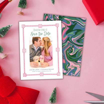 A white wedding invitation card with a photo of a couple and the text "Deck the Hell of Save the Date" is placed on a pink background, surrounded by Christmas decorations and a red gift box.