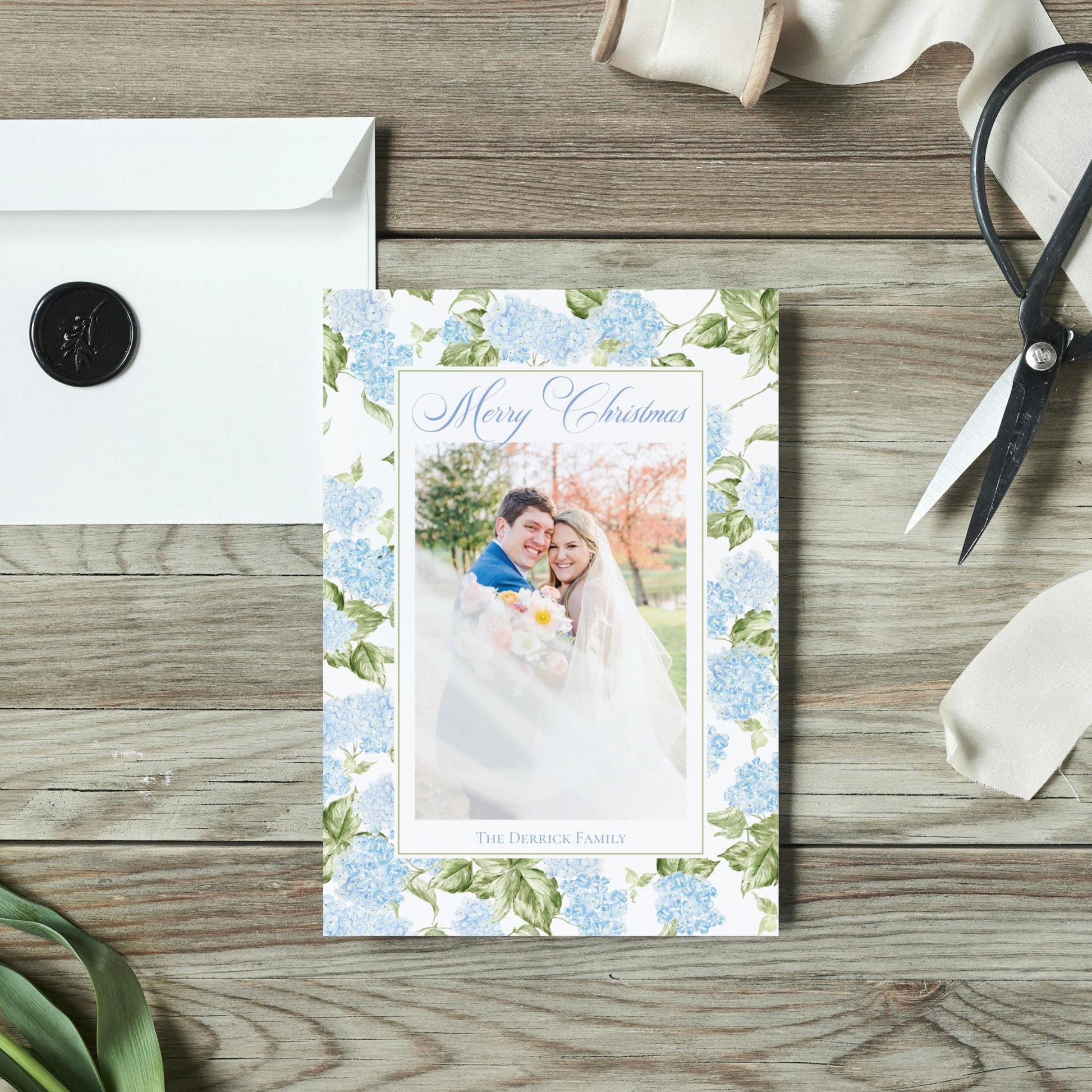A wedding card with a couple in a floral design, placed on a wooden surface alongside a white envelope, a pair of scissors, and some ribbon.