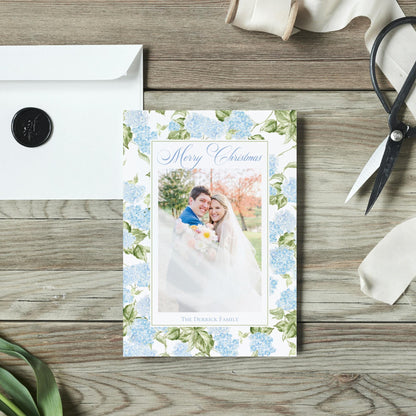 A wedding card with a couple in a floral design, placed on a wooden surface alongside a white envelope, a pair of scissors, and some ribbon.