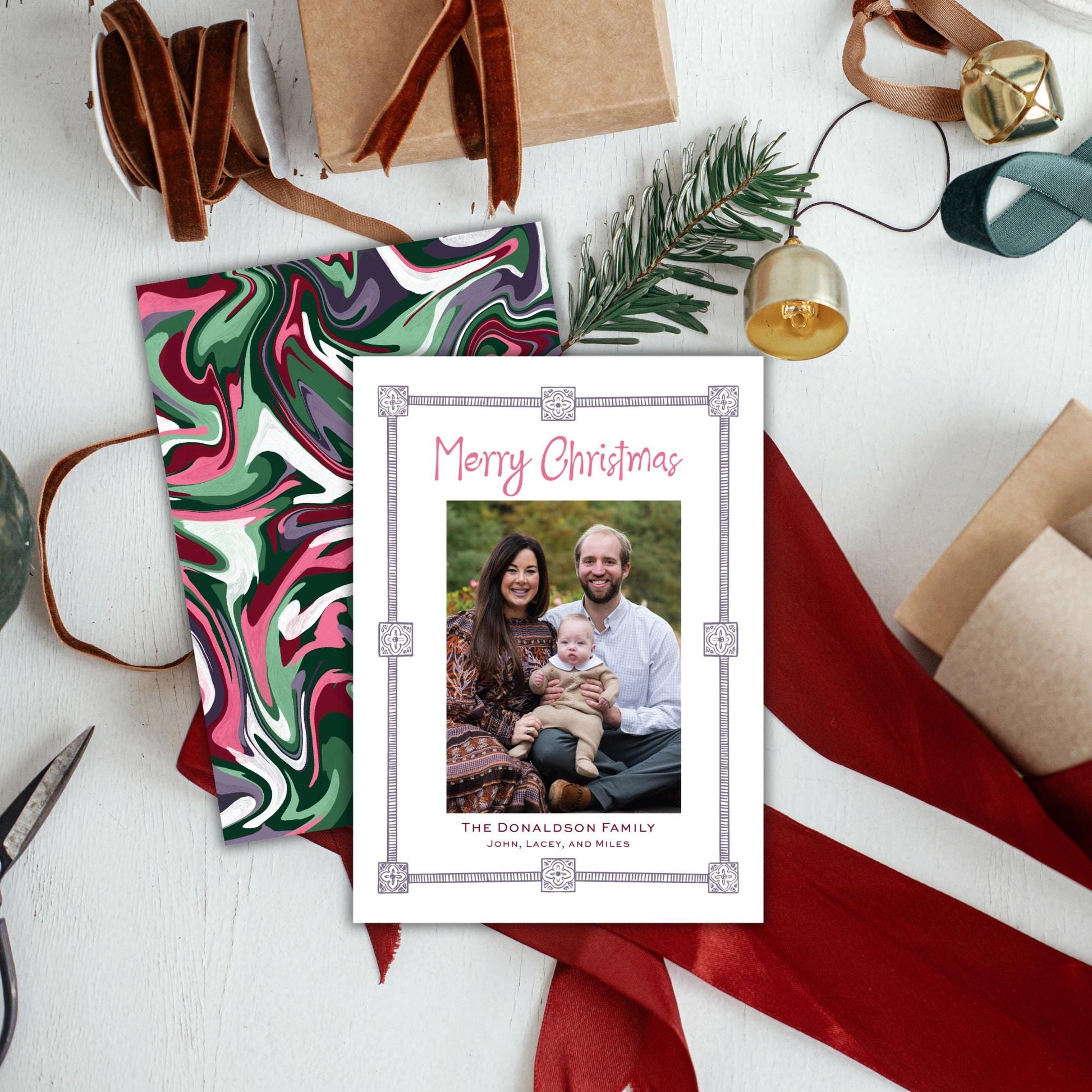 A Christmas card with a family photo is displayed on a table surrounded by holiday decorations, including a Christmas tree, ribbons, and other festive items.