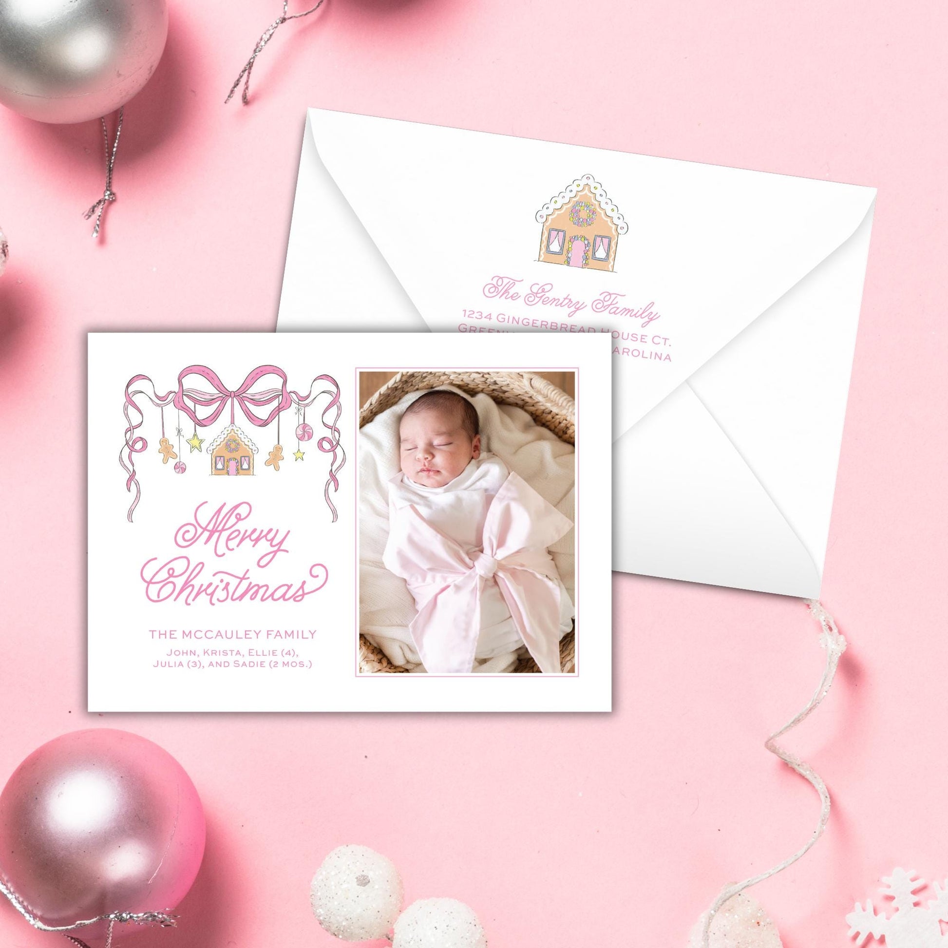 A white envelope with a photo of a sleeping baby inside, along with a pink bow and a Christmas-themed design. The envelope is placed on a pink background with some ornaments and a snowflake decoration.
