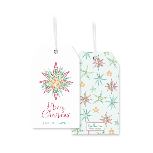 two white tags with Christmas-themed designs hanging from strings. The tag on the left has a star-shaped design with a floral pattern, and the tag on the right has a star-shaped design with a snowflake pattern. Both tags display the text "Merry Christmas" and "Love, The Paynes" in a festive font.