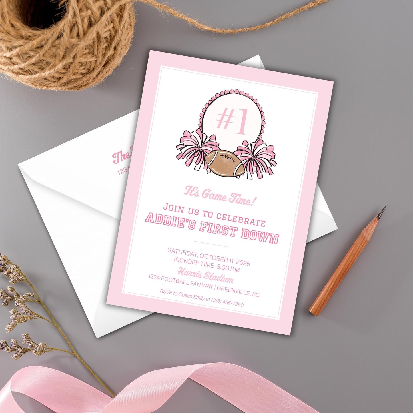 a pink invitation card with a football graphic and text, placed on a gray surface alongside a brown string, a pencil, and some greenery.