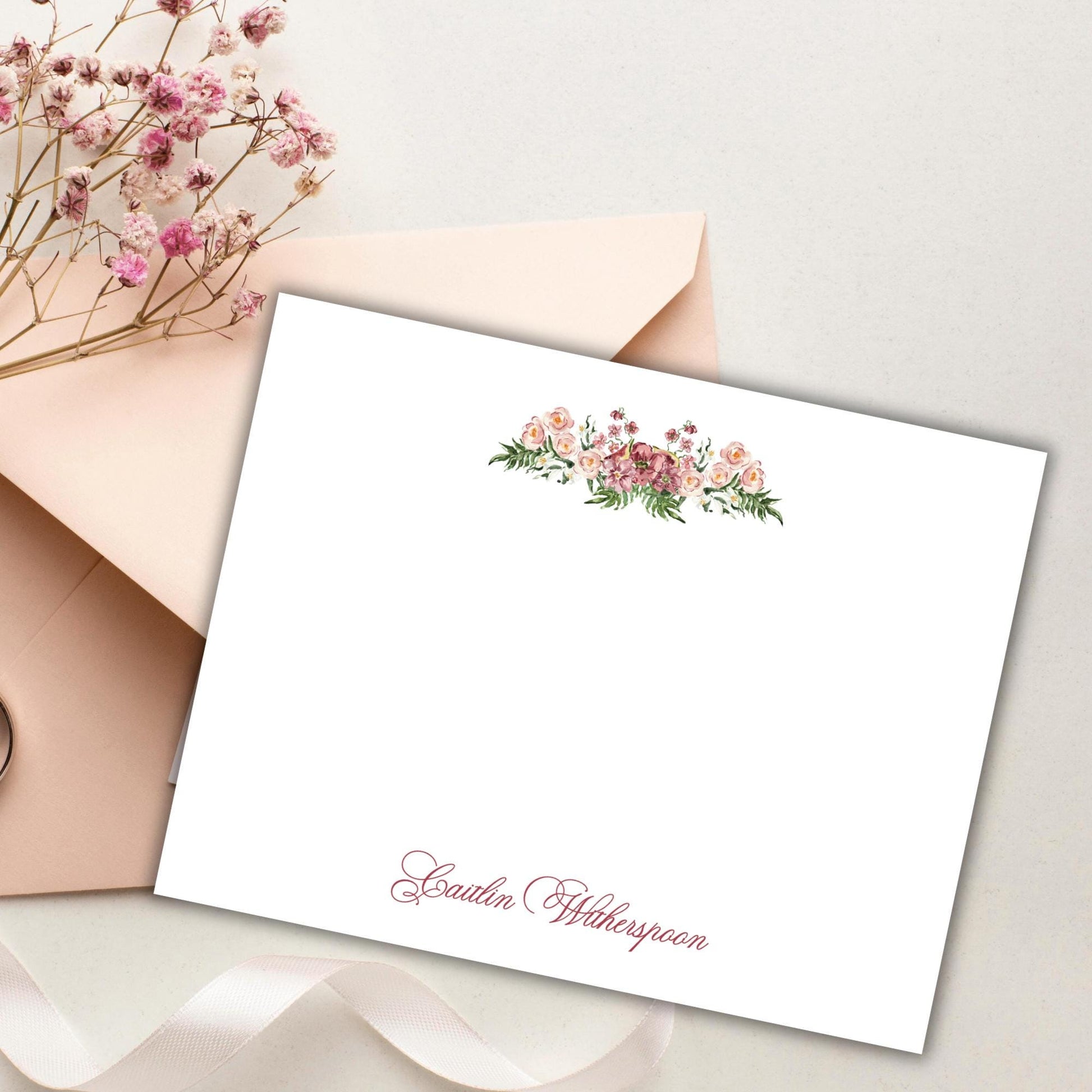 a white card with a floral design and the name "Gailin Wheatspoon" written on it, placed on a surface with a pink envelope and some dried flowers.