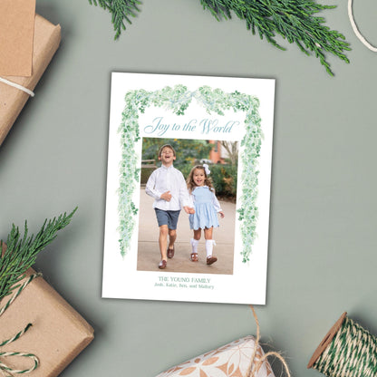 A white card with a photo of two children, surrounded by greenery and holiday decorations, including wrapped gifts and sprigs of pine.