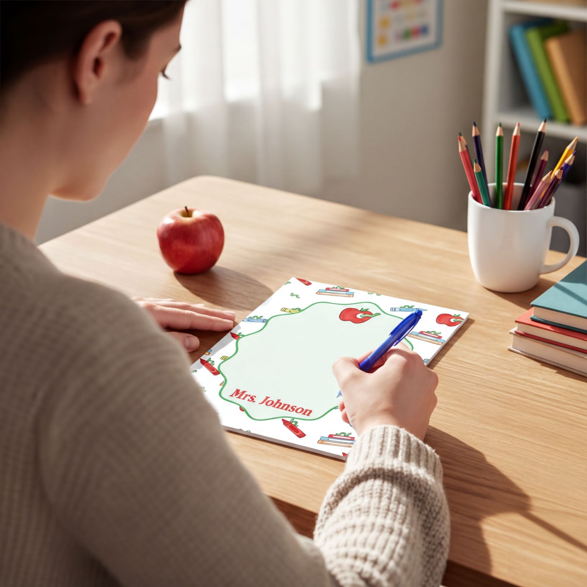 A person is sitting at a desk, writing on a piece of paper with a blue pen. There is a cup of colored pencils and an apple on the desk.