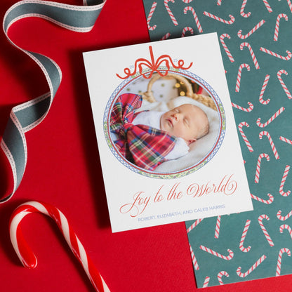 A white card with a photo of a sleeping baby and the text "Joy to the World" is placed on a red surface, accompanied by a red and white striped ribbon and a candy cane.