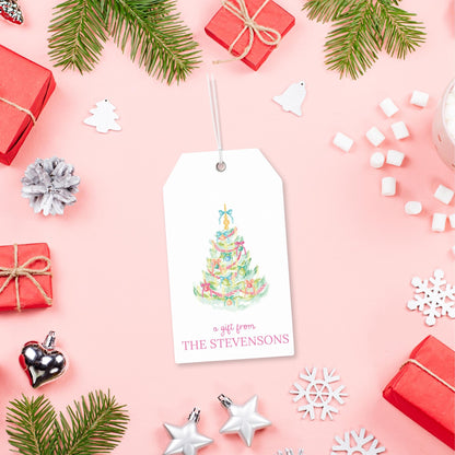 A white gift tag with a Christmas tree illustration and the text "a gift from THE STEVENSONS" is displayed on a pink background, surrounded by various Christmas decorations such as pine branches, red gift boxes, and snowflakes.