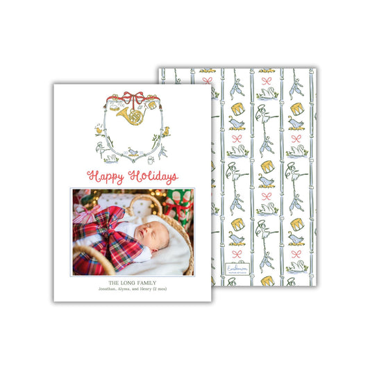 A holiday card featuring a baby sleeping in a basket, with a wreath and a patterned background.