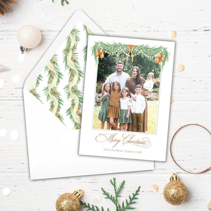A family photo is displayed on a white card, surrounded by Christmas decorations such as ornaments, pine cones, and greenery.
