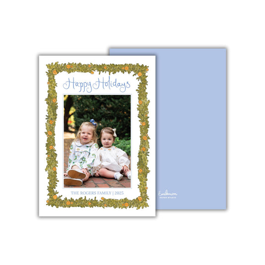 A holiday card featuring two young girls sitting on a brick path, surrounded by a decorative border of leaves and oranges. The card has a blue background and the text &quot;Happy Holidays&quot; written in a decorative font.
