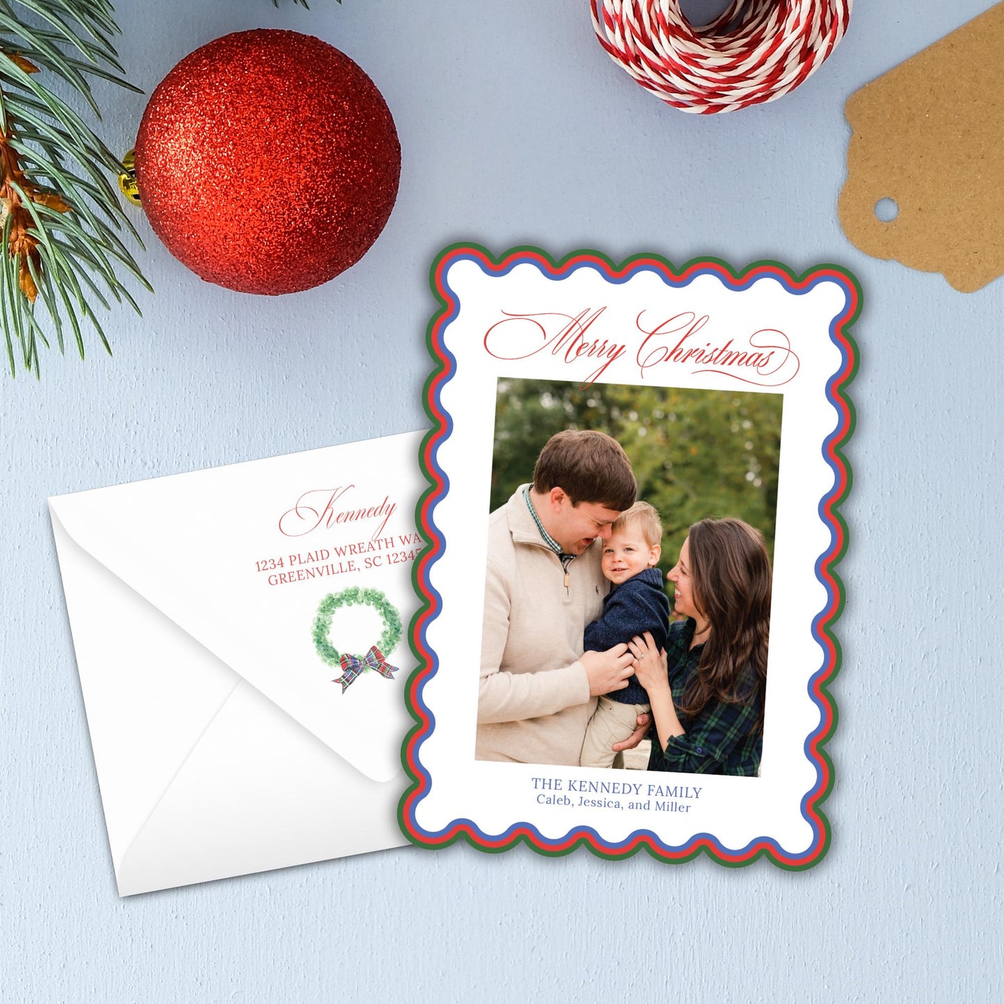 A Christmas card with a family photo of a man, woman, and child, along with a wreath and a red ornament, all placed on a blue background.
