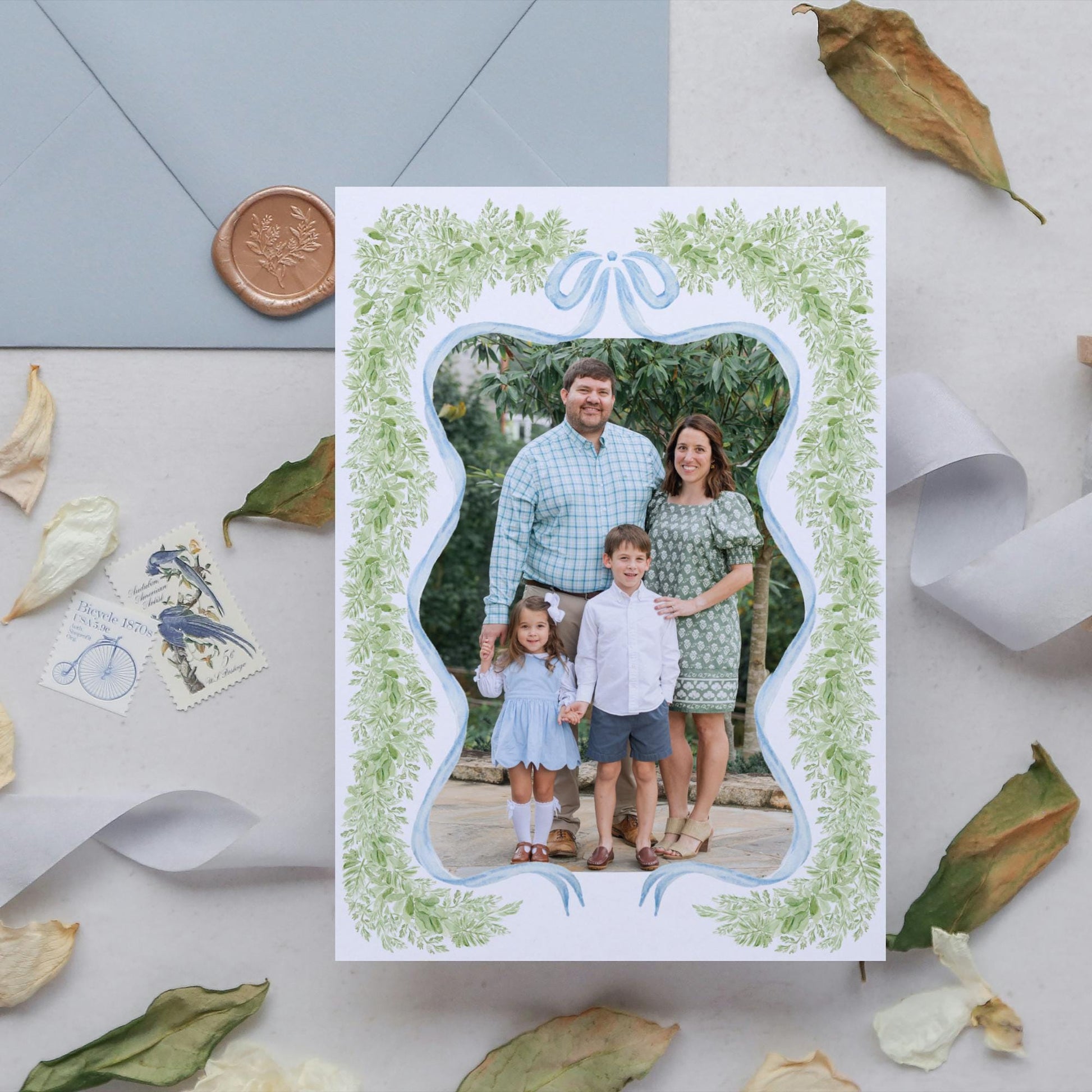 A family portrait of a man, woman, and two children, framed in a decorative border with green leaves, is displayed on a white card. The card is surrounded by autumn leaves and postage stamps, creating a seasonal and nostalgic atmosphere.