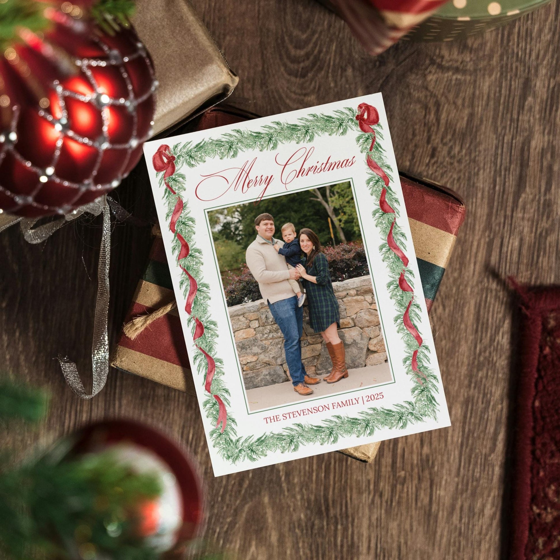 A Christmas card with a family photo is placed on a wooden surface, surrounded by wrapped gifts and ornaments.