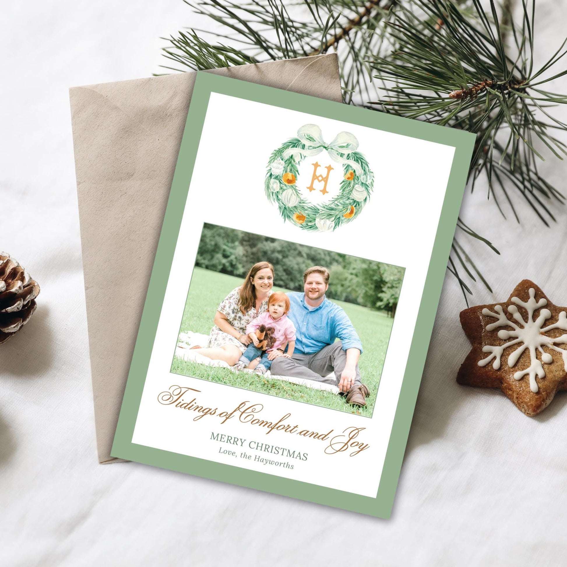 A Christmas card with a family photo and a wreath, along with some pine cones and a star-shaped cookie, all placed on a white surface.