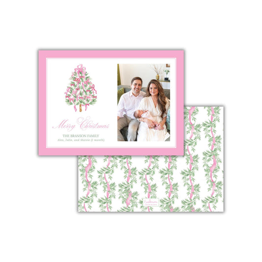 A pink and white Christmas card featuring a family photo of a man, woman, and baby. The card has a floral pattern and the text &quot;Merry Christmas&quot; and &quot;THE BRANSON FAMILY Alex, Julie, and Moisie (1 month)&quot; printed on it.
