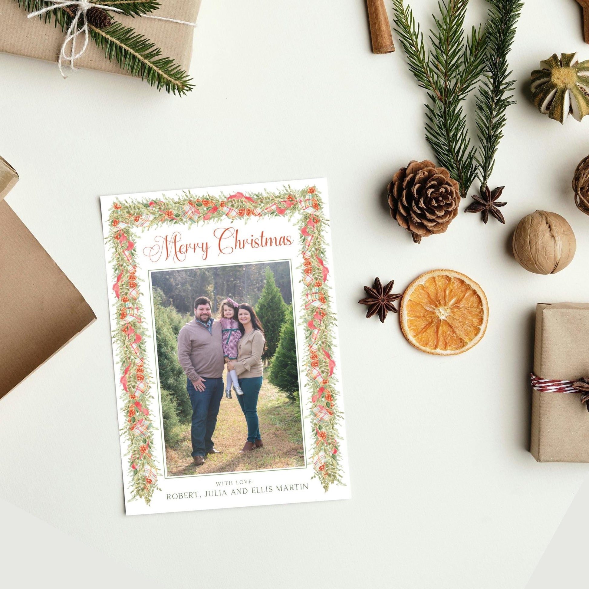 A Christmas card with a family photo is displayed on a white surface, surrounded by holiday decorations such as pine branches, pine cones, and an orange slice.